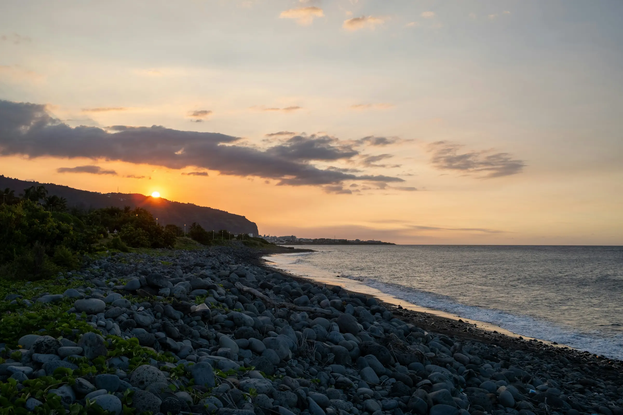 The sun sets behind a pebbled and green coastline on Reunion Island.